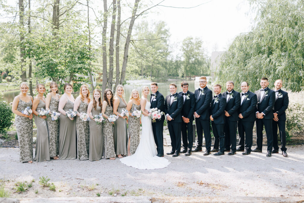 Wedding party with bridesmaids in sage green floral dresses infront of green spring trees and a pond at Swan Lake wedding venue