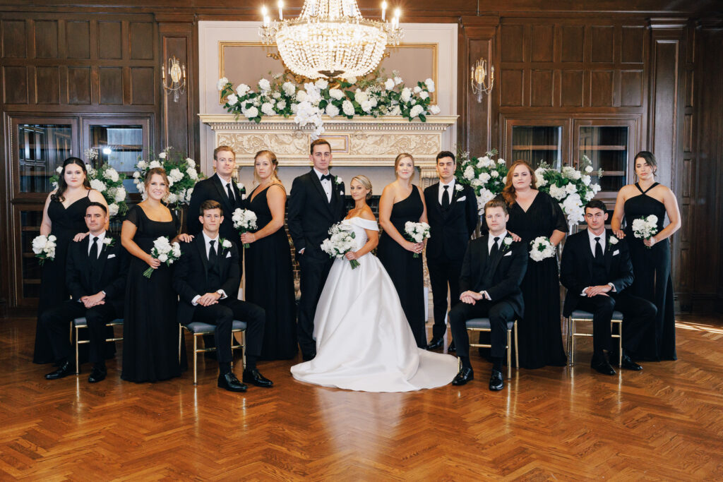 bridal party standing infront of formal fireplace covered in white roses with chandelier at the athletic club wedding venue