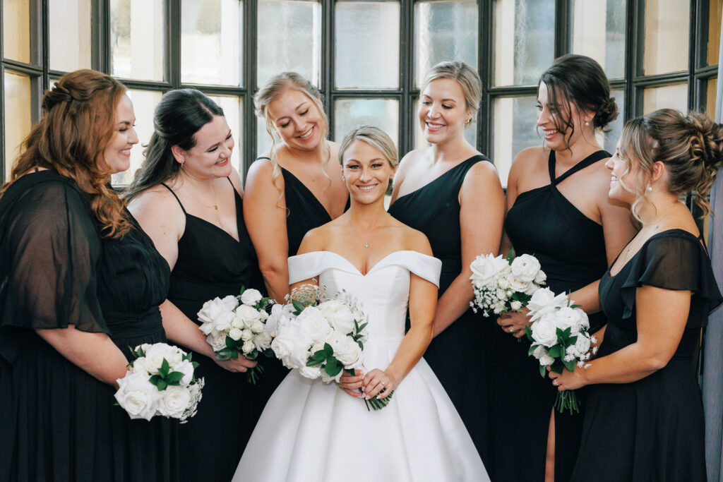 bride with bridesmaids in black gowns standing infront of large window at the Athletic Club Wedding venue
