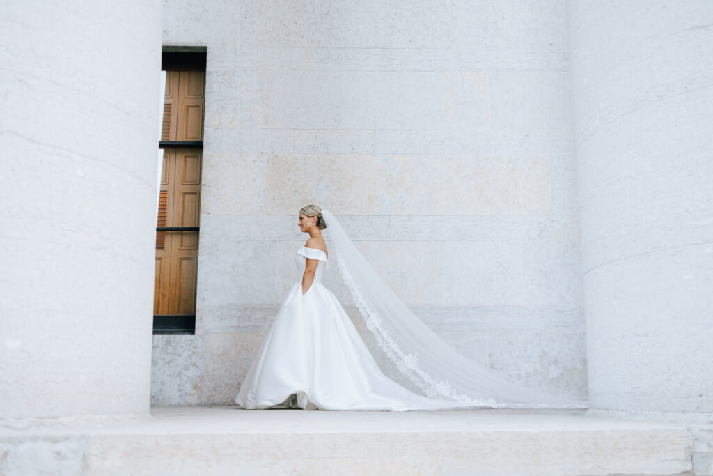 brides stands infront of marble wall with large pillars with long lace veil