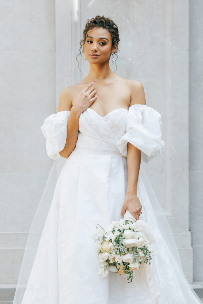 Bride standing in of marble wall with curly hair updo and wedding gown with puff sleeves holding bouquet at the Columbus Museum of Art wedding venue